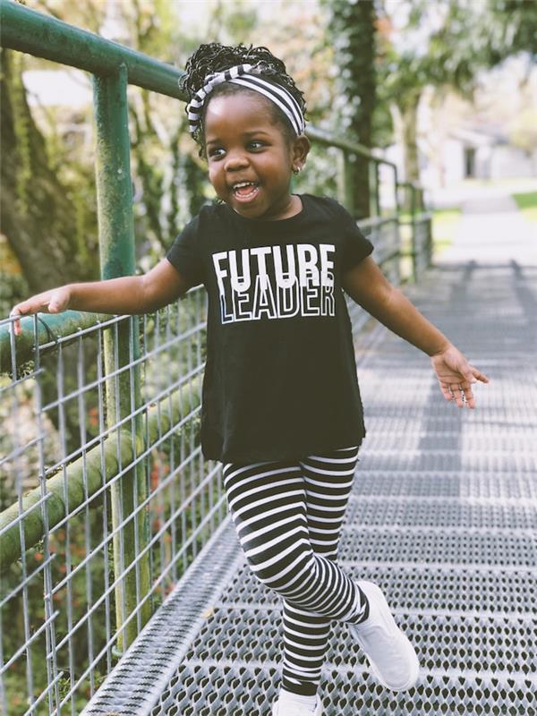 Rozkošné malé dievčatko v čiernobielom oblečení s gumičkou do vlasov, jednoduchým účesom s chrumkavými vlasmi a usmievavým dievčaťom