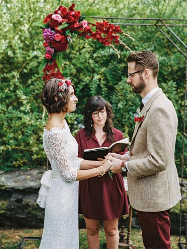 عروس وعريس يقولان نعم أمام قوس به ورود حمراء ووردية ، تسريحة شعر زفاف ذات شعر قصير ، تاج عروس بالورود والأوراق