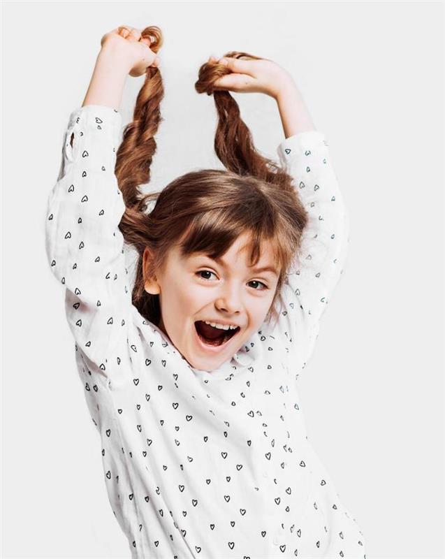 Dva vrkoče na dlhých vlasoch usmievavého dievčaťa v bielej košeli so srdiečkami, ako urobiť účes pre malé dievčatko, jednoduchý návod na účes