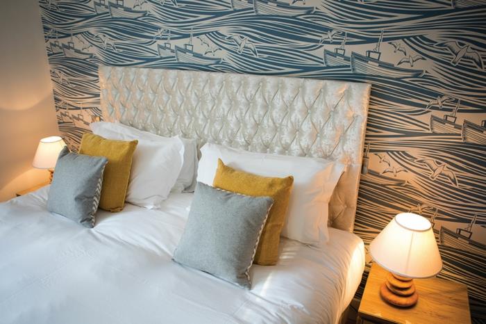 drevený kus nábytku, originálne čelo postele, potiahnuté saténovou tkaninou v dúhových farbách s prešívanými efektmi, stena pokrytá tapetami v modro -bielych farbách, morské vzory s veľkými vlnami a malými člnmi, ktoré sa motajú