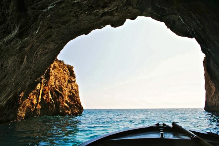 grottans utgång med en liten fiskebåt, molnfri himmel, blått vatten Grotta azzurra, bruna och grå stenar