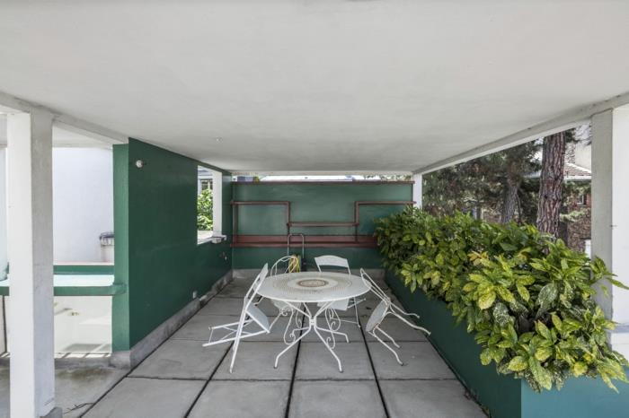 vnútorná zelená stena, oddeľovač zeleného priestoru, rastliny v zelenej a žltej farbe, moderná veranda, biely kovový záhradný nábytok, štyri skladacie stoličky a okrúhly stôl