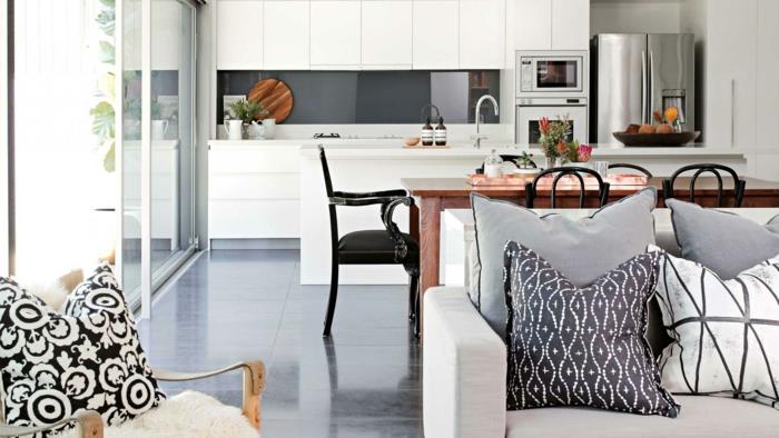 malá otvorená kuchyňa v bielej farbe so sivým splashbackom v lesklom prevedení, drevený stôl a čierne kovové stoličky, sivá dláždená podlaha, sivá sedačka a sivé, čiernobiele vankúše