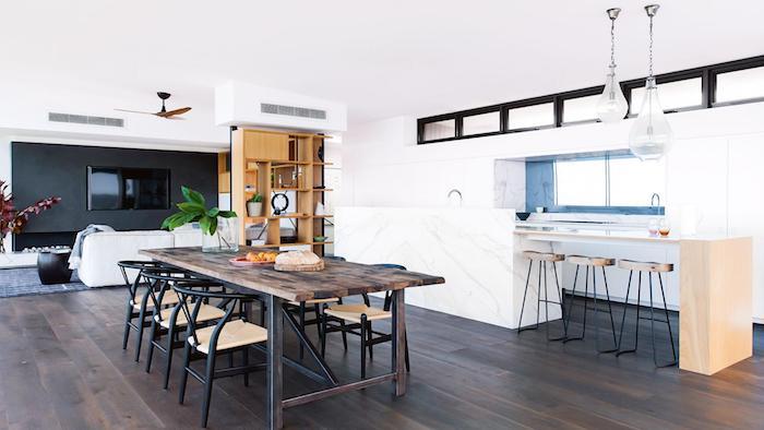 kuchyňa otvorená na obývaciu izbu a jedáleň, drevený ostrov so stoličkami, modrý farebný splashback, hnedé parkety, drevený stôl a čierne kovové stoličky, akcentovaná stena s TV v obývačke a rozbitá biela pohovka