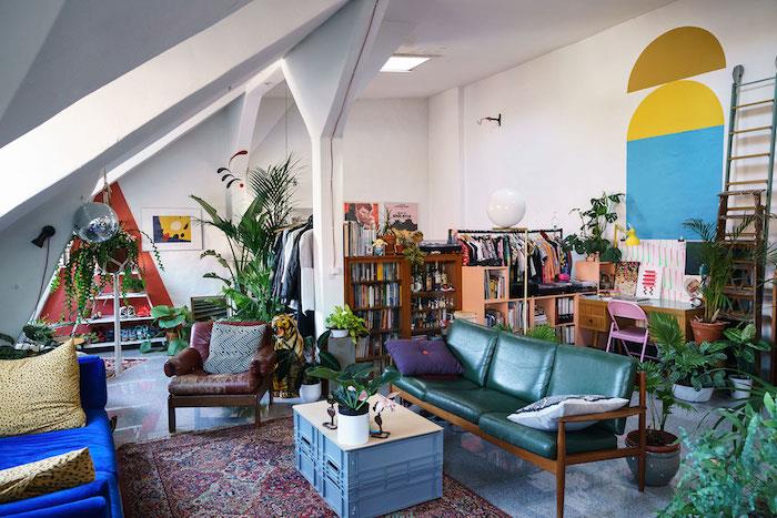 nápad, ako zariadiť štúdio, drevenú pohovku so zelenými vankúšmi a ďalšou modrou sedačkou, originálny kancelársky priestor na uskladnenie obuvi, orientálny koberec, dekor džungle vo farbách a rastlinách