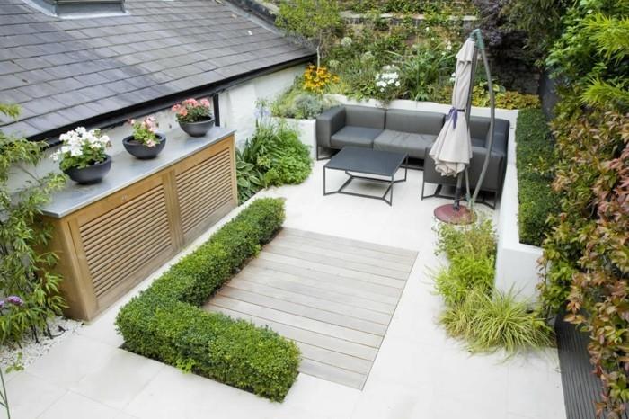 trädgårdsmöbelidé, grå soffa och bord på en Zen -terrass, träbeklädnad, kakel, parasoll, boxwood, buskar och träd, grus på kanterna