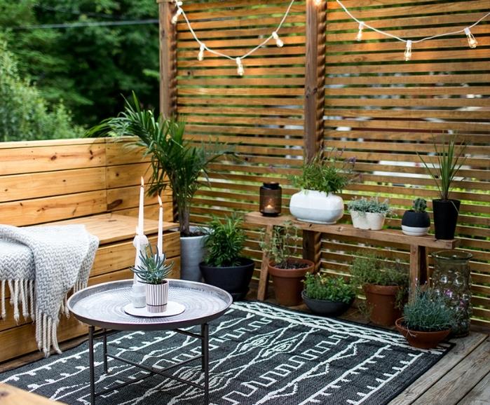 deco träterrass, träsoffa och brisvy, svart matta med vita geometriska mönster, designer soffbord, grå pläd, gröna växter, ljus krans