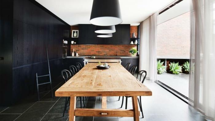 vybaviť malú kuchyňu, čiernu fasádu kuchynskej linky, čierne závesné svetlá nad dreveným stolom, obklopené kovovými stoličkami, sivú dláždenú podlahu a murovanú kuchyňu