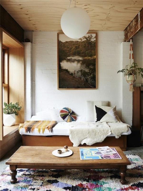 سجادة منسوجة متعددة الألوان ، داخل غرفة معيشة صغيرة ، أريكة خشبية مغطاة بأغطية وبطانيات ، تصميمات بسيطة لغرفة المعيشة ، طاولة قهوة عتيقة منخفضة