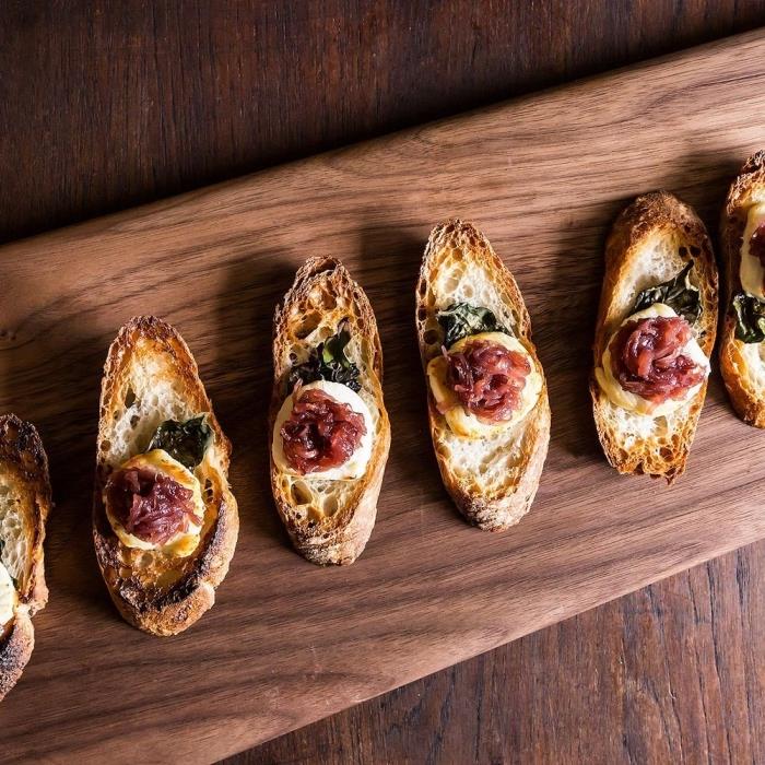 plátky toastu sprevádzané kozím syrom a cibuľovým konfitom umiestnené na drevenej prezentačnej doske, aperitívový toast s kozím syrom a cibuľovým konfitom