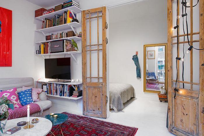 sovrum öppet för ett vardagsrum med grå soffa dekorerad med färgglada kuddar, röd orientalisk matta, marmorsoffabord, trädörrar med gamla fönsterluckor, bohemisk chic studio deco