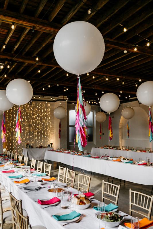 biele héliové balóniky s ozdobou z kúskov farebného papiera nad stolom s bielym obrusom zdobeným viacfarebnými obrúskami