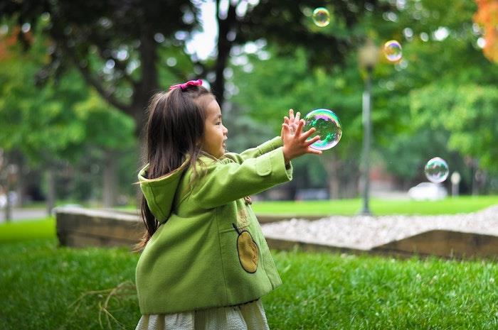 Dievčatko sa hrá s mydlovými balónikmi, účes malé dievčatko, účes vrkoč, ľahký návod na účes, zelený kabát, zelená záhradná jar