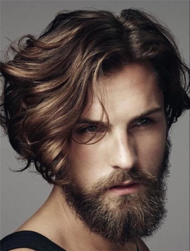 bradatý muž s vlnitými hnedými vlasmi, na jednej strane ostrihaný nakrátko a na druhej ponechaný dlhší, prameň vlasov mu padá na oko