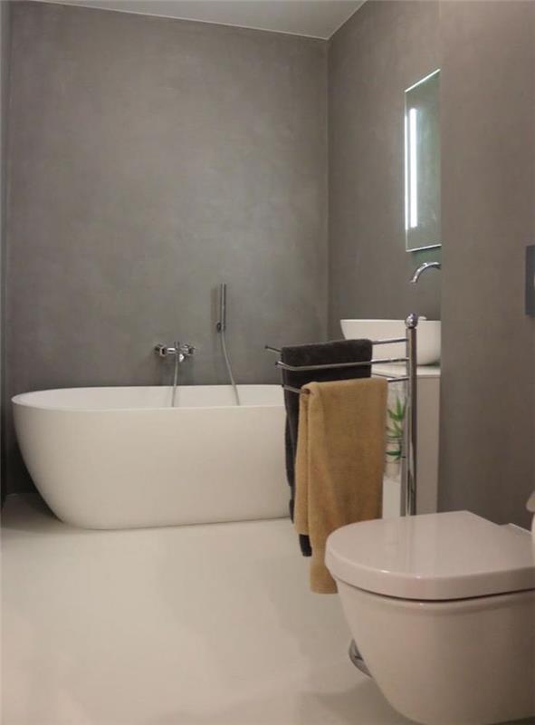 Zrekonštruovaná kúpeľňa v modernom štýle so sivou stenou z voskovaného betónu, bielou dlažbou a oválnou voľne stojacou vaňou
