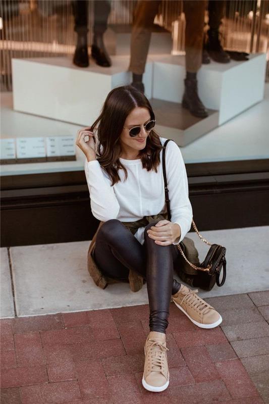امرأة تجلس في الشارع ، اتجاهات الموضة الحالية ، ترتدي لباس ضيق من الجلد الأسود وبلوزة بيضاء ، أحذية رياضية بيج وحقيبة سوداء