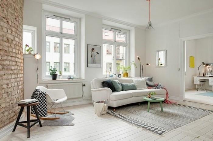 غرفة معيشة على الطراز الاسكندنافي باللون الأبيض ، مع لمسات من الطوب على الحائط ، وتحتوي على أريكة بيضاء ، مع العديد من الوسائد ، وسجادة منقوشة باللون الرمادي ، وكراسي وطاولة قهوة فيروزية ، وأفكار لتجهيز الغرفة