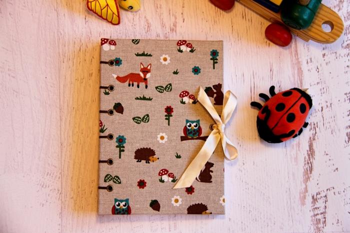 denník pre deti potiahnutý béžovou ľanovou tkaninou s motívmi lesných zvierat, prítulná lienka, stôl s bielou drevenou doskou, bábätko