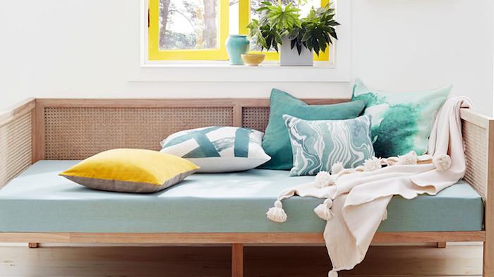 drevená sedačka s matracmi v seladonových farbách, zdobená vankúšmi, modrá, zelená a žltá, svetlo sivý poťah, okná so žltými rámami, stena na bielom pozadí