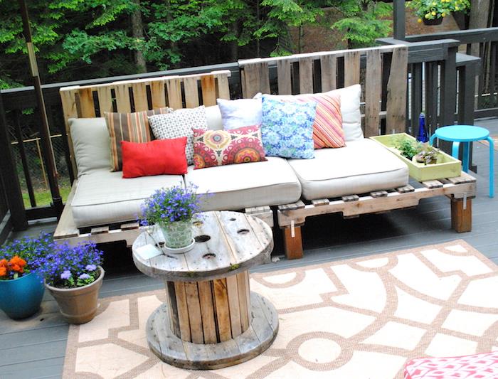 príklad záhradného nábytku z paliet s drevenou paletovou sedačkou so sivým vankúšom na sedenie a farebnými ozdobnými vankúšmi, navijakom na stôl, ozdobou terasy v kvetináči