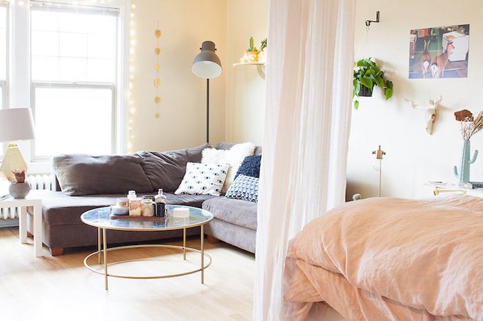sovrumsdelning i vardagsrummet med en transparent slöja-gardin, grå soffa med dekorativa kuddar, laxlinne på sängen, ljus parkett, glas- och mässingsbord