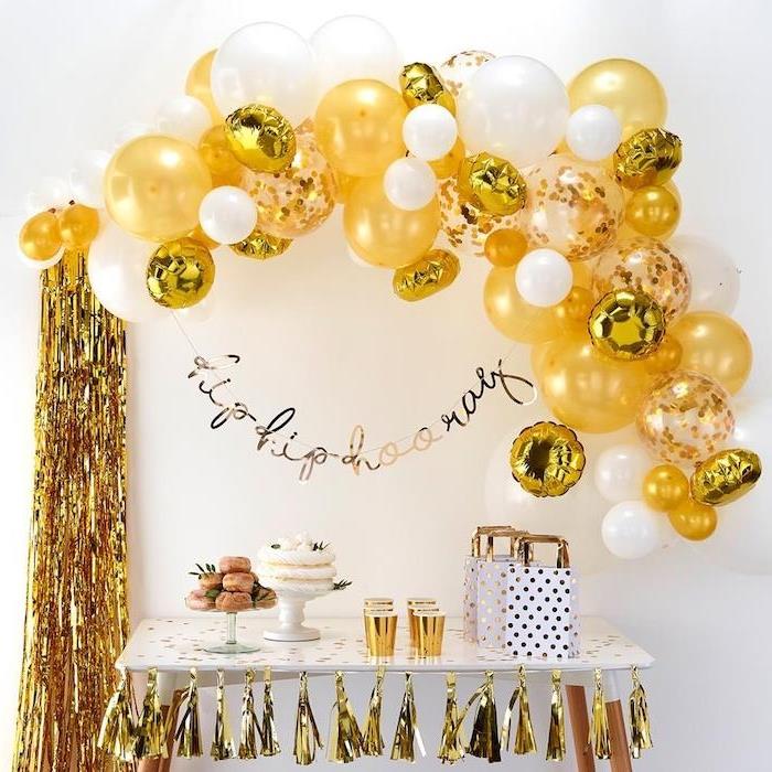 båge av guld- och vita ballonger på vit väggbakgrund, krans av pomponger med gyllene fransar, festgodisbar med munkar och kakor