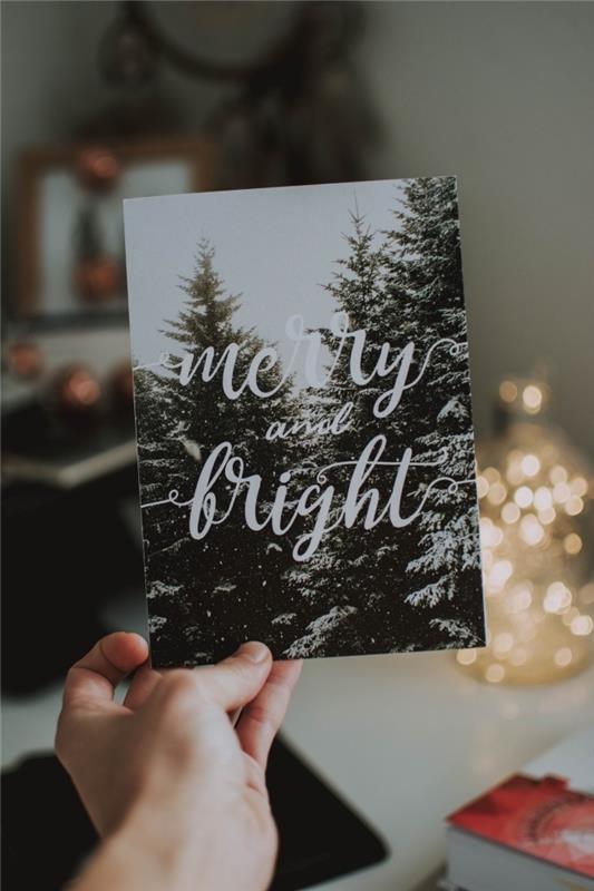foto av ett julkort med en snöig landskapsbild och Happy Holidays bokstäver framför en mini vit julgransdekoration på ett vitt skrivbord