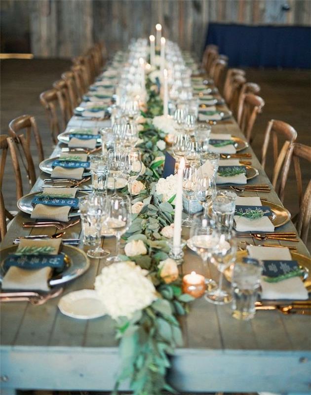 stredobod s vavrínovým vencom a kvetmi na stole zo surového dreva, zlaté taniere a príbory, jedálny lístok napísaný na čiernom papieri, ozdobné sviečky, rustikálna výzdoba svadobného stola