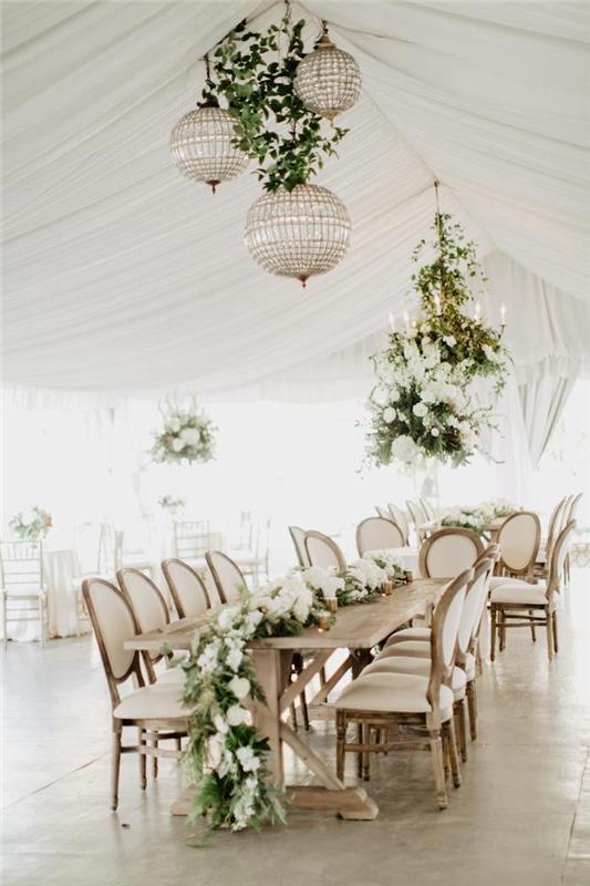 zlatý a sklenený luster a vegetácia zavesené nad stolom zo svetlého dreva zdobeného girlandou z listov a bielych kvetov, bielym stropným závesom