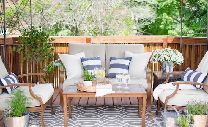trädgårdsmöbelidé i komposit med soffa, fåtöljer och bord i trä, vita och grå sittdynor, grå, blå och vita dekorativa kuddar, gröna växter