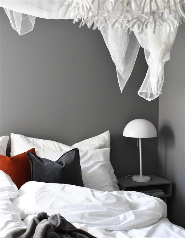 výzdoba hlavnej spálne so sivými stenami a sivou, bielou a červenou posteľnou bielizňou, jednoduchá dizajnová nočná lampa