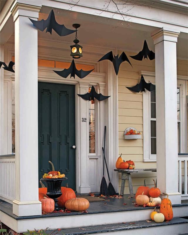 halloween dekoration av verandan, beige och vitt husfasad med mörkgrön dörr, dekoration med orange pumpor och svarta pappersfladdermöss