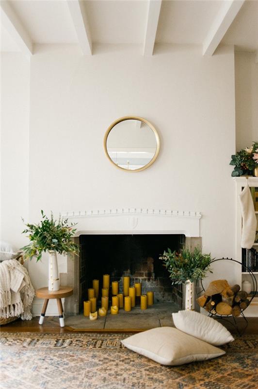 مدفأة مدمجة في الجدار الأبيض مع رف منحوت بسيط مع شموع صفراء في الموقد ومرآة دائرية ذهبية عتيقة