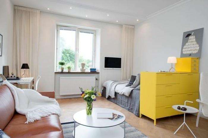 أريكة جلدية باللون البني ، مع غطاء أبيض ، بالقرب من طاولة قهوة بيضاء مستديرة ، داخل غرفة بأرضية خشبية ، وسرير وخزانة ذات أدراج صفراء