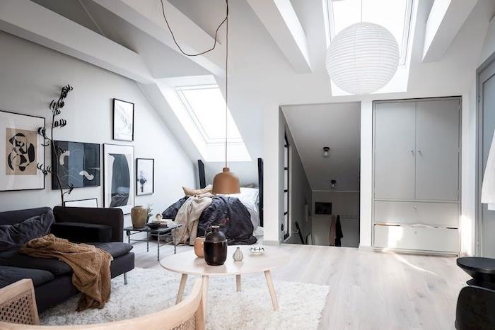 sluttande säng på ett vindsutrymme inrett i skandinavisk deco -stil, koksgrå soffa, vit matta, bord i skandinaviskt trä, parkett i ljust trä, vita synliga balkar