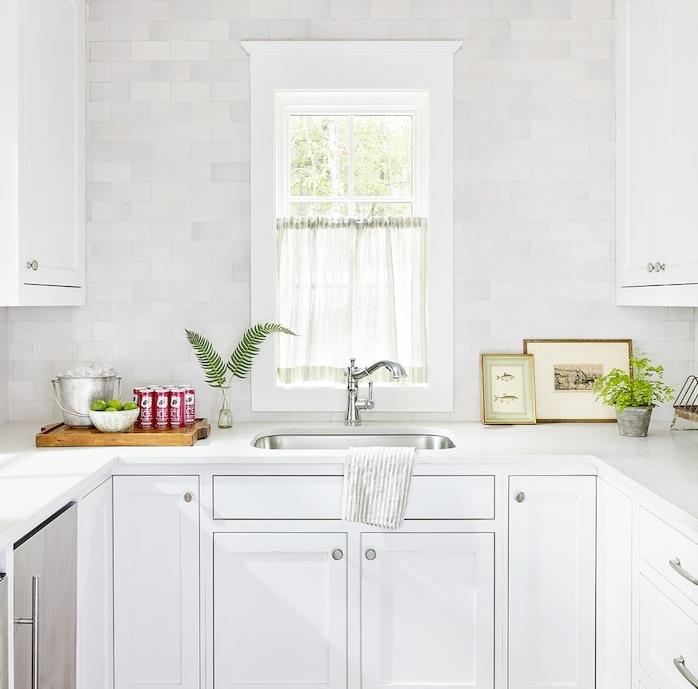 litet vitt utrustat kök, köksskåp och vita väggplattor, rostfria detaljer, växter, skärbräda