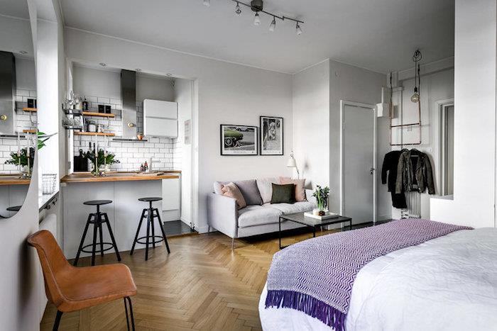 exempel på funktionell studiolayout med säng framför ett vitt och grått pentry med liten bar, matbord, vitt tunnelbana kakel och vardagsrum med grå soffa och svart soffbord