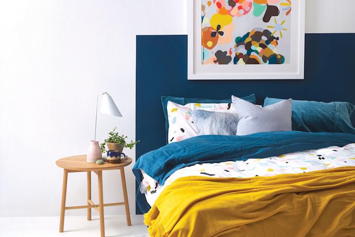 Dekorácia spálne pre dospelých, bielo namaľované steny a modré čelo postele, drevený nočný stolík, modrá, žltá a biela bielizeň, dekorácia rámu s abstraktným obrazom