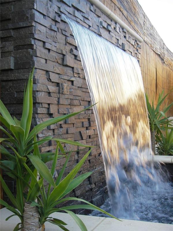 ozdobte vonkajšiu stenu obdĺžnikovými čiernymi kameňmi usporiadanými asymetricky, fontána, záhradný vodný lúč, vodopád so zelenými rastlinami na oboch stranách