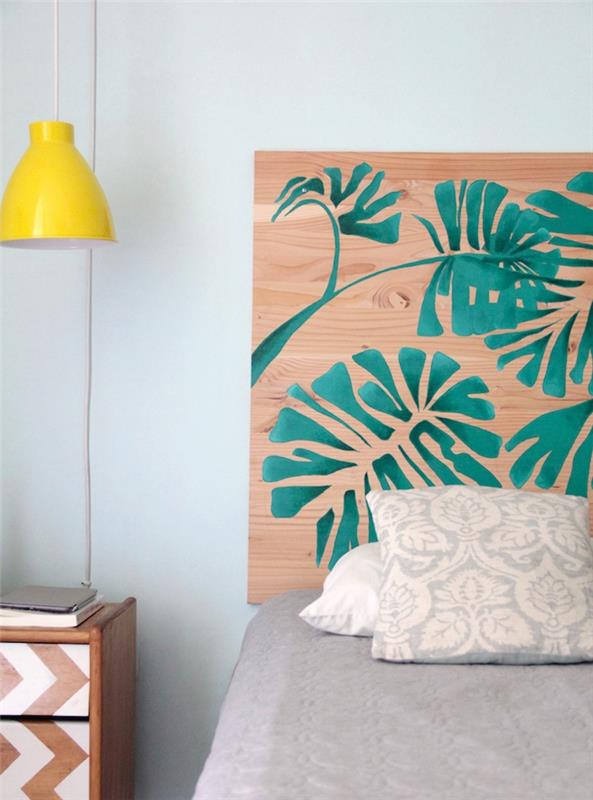Huvudgavel av plywood dekorerad med gröna palmblad, grått sänglinne, sängbord i trä dekorerad med vit färg
