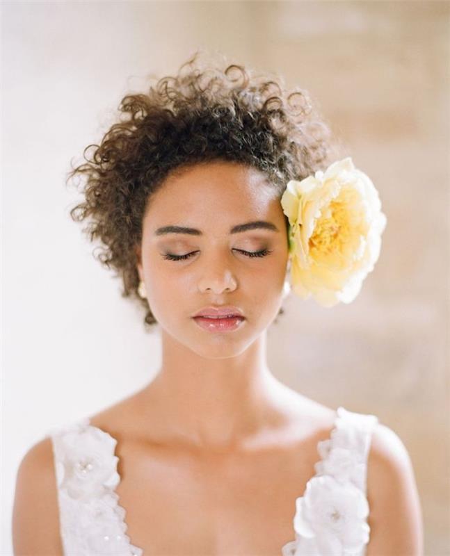 تصفيفة الشعر الأفرو امرأة مع الشعر المجعد اكسسوارت مع وردة صفراء كبيرة على جانب الرأس ، فستان الزفاف الأزهار البيضاء
