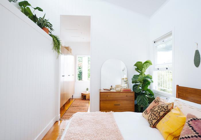 bohémsky elegantný dekor spálne s drevenou posteľou s bielym ľanom a ružovým plédom, farebnými vankúšmi, drevenou komodou, bielymi stenami, zelenou rastlinou v kvetináči a vysokou vegetáciou