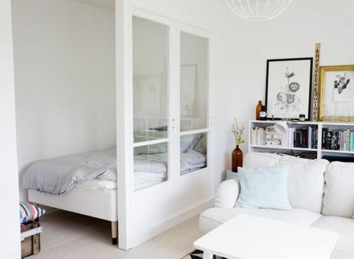 منطقة نوم مع سرير صغير ، نصف منفصلة عن منطقة غرفة المعيشة ، من خلال جدار جزئي أبيض ، مع إدخالات زجاجية ، وتصميم شقة ، وأريكة بيضاء اللون وأرفف كتب في مكان قريب