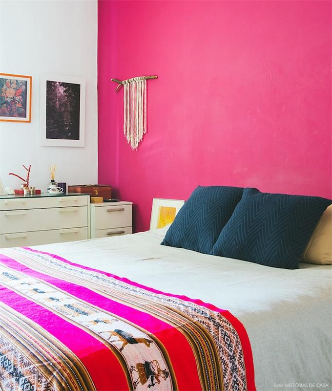 obraz do spálne pre dospelých s fuchsiovou ružovou akcentovanou stenou, čiernymi vankúšmi, orientálnym vzorom prikrývky na posteľ, bielou komodou, dekoráciou steny macrame