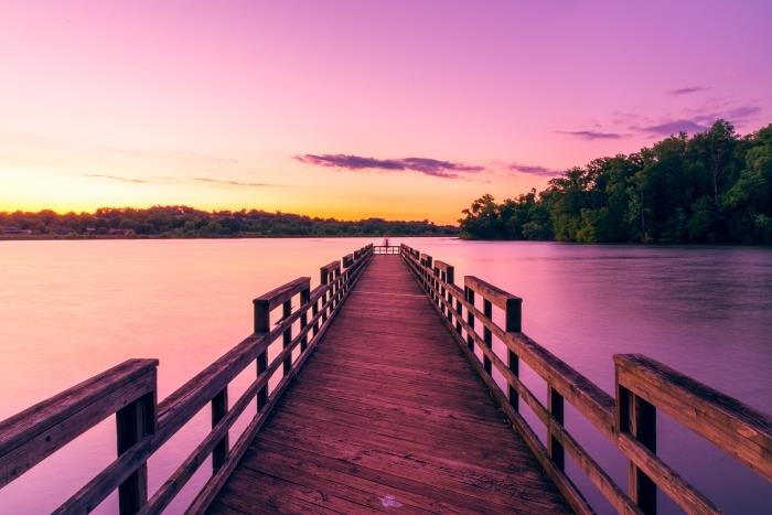 drevený most cez jazero so stromovým lesom a oblohou v ružových a žltých farbách osvetlených slnečnými lúčmi, pozadie tapety pri východe slnka
