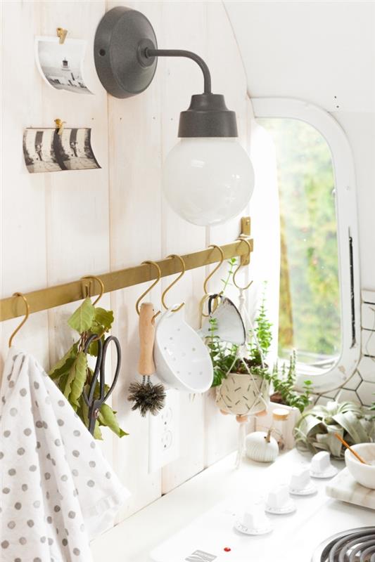 mosadzná tyč na držanie kuchynského náčinia s malými háčikmi inštalovanými na stene za sporákom, doplnená starým nástenným svetlom, úložné riešenia na optimalizáciu a usporiadanie malej kuchyne