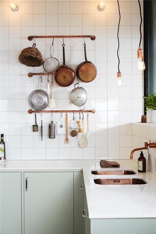dekoratívny tip na aranžovanie funkčnej kuchyne v štýle bistro, vintage škandinávska kuchyňa v bielej a mandľovo zelenej farbe s medenými akcentmi, držiaky na kuchynské pomôcky s medenými rúrkami pripevnené k stene