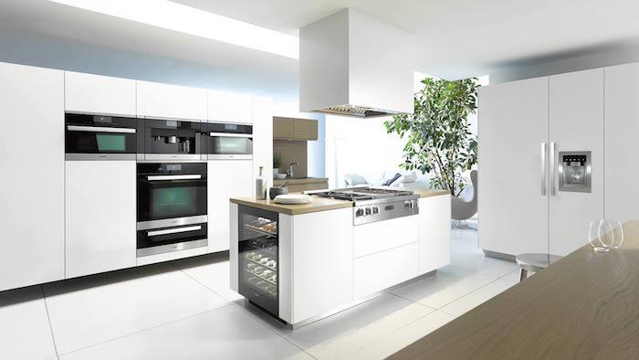 vitt utrustat kök med vitt skåp och ö och vitvaror i svart och rostfritt stål, vita golvplattor, grön växt