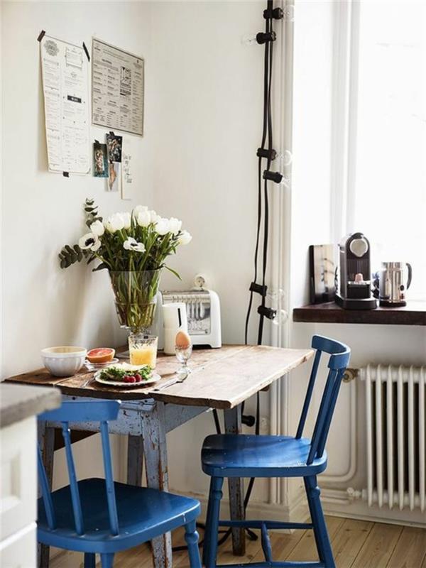 dekorácia kačacia modrá, nábytok z kačacej modrej, malý štvorcový hrubý drevený stôl, biele steny, svetlá girlanda s žiarovkami okolo okna