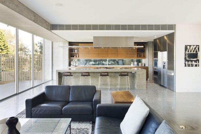 model kuchyne otvorený do obývačky, drevená fasáda kuchynskej linky a sivý bar s drevenými a kovovými stoličkami, náter, betónový efekt, sivá sedačka, sklenený stôl